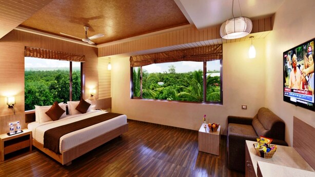 A spacious bedroom with a large bed, a sofa, and a TV. It features wooden flooring, large windows with a view of greenery, and a ceiling fan. - Sarovar Portico Gir (Mendarda)