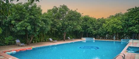 A rectangular swimming pool with clear blue water, surrounded by lush green trees and a tiled deck.