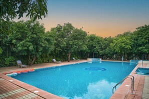 A rectangular swimming pool with clear blue water, surrounded by lush green trees and a tiled deck. - Sarovar Portico Gir (Mendarda)