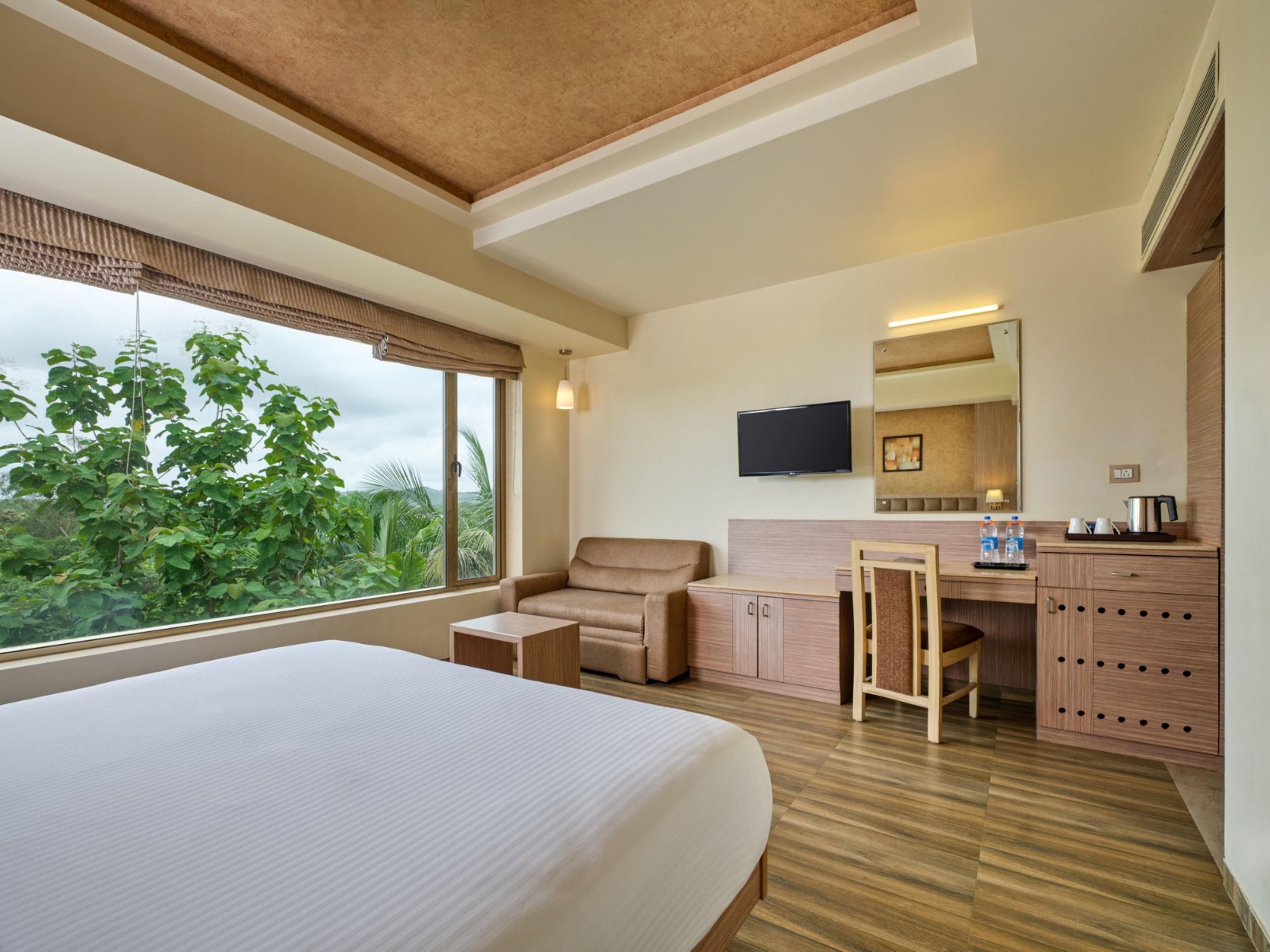 Habitación de hotel con cama, escritorio, silla, televisor y ventana con vistas a zonas verdes.