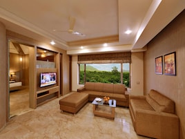 A modern living room with a sofa, ottoman, and coffee table. There is a television on a wooden stand, and a window with a view of greenery.