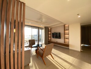A modern living room with a flat-screen TV, a dining area with a table and chairs, and a large window offering a view. - Sarovar Portico Gir (Mendarda)