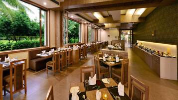 A restaurant with multiple dining tables and booths, large windows offering a view of greenery, and a buffet counter with various food items.