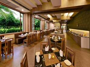 A restaurant with multiple dining tables and booths, large windows offering a view of greenery, and a buffet counter with various food items. - Sarovar Portico Gir (Mendarda)