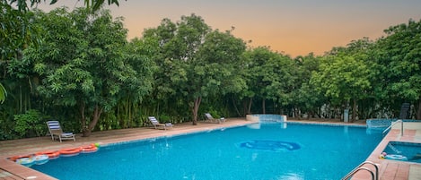 Una piscina rectangular con borde de azulejos, rodeada de frondosos árboles verdes.
