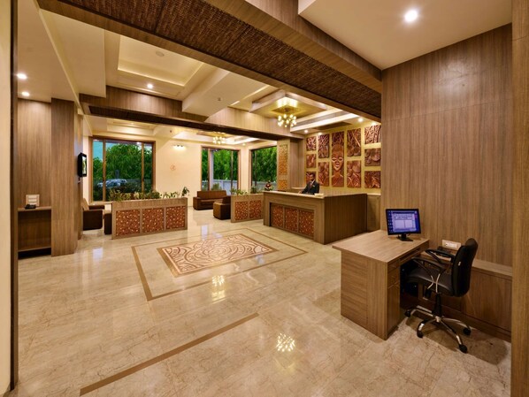 A modern reception area with a wooden reception desk, a computer, and a seating area with a patterned rug. - Sarovar Portico Gir (Mendarda)