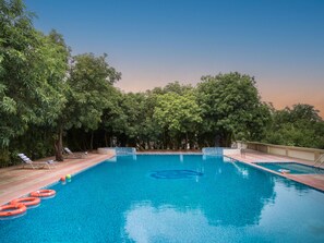 A swimming pool with clear blue water, surrounded by trees and shaded by a roof. - Sarovar Portico Gir (Mendarda)