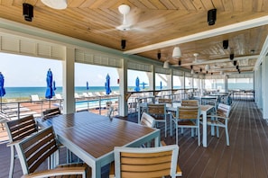 Een overdekt terras met houten tafels en stoelen, met uitzicht op een zwembad en strand.