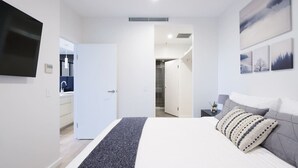 A modern bedroom with a large bed, a flat-screen TV, a small bedside table, and a door leading to a bathroom. - Sydney CBD 2-Bedroom Apartment with Hyde Park Views - Sleeps 6 (Sydney)