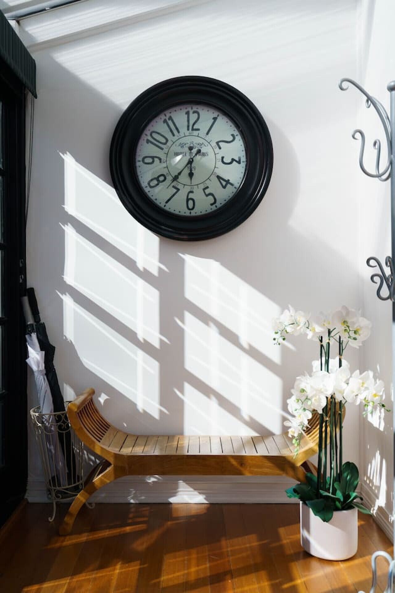 A wall-mounted clock, a wooden bench, a white orchid vase, and a metal umbrella stand.