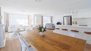 Uma sala de estar e jantar moderna, com conceito aberto, mesa de jantar de madeira, mobiliário branco e uma cozinha com armários e eletrodomésticos brancos.