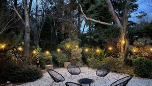 Uma área externa com assentos, cadeiras e uma mesa, rodeada por cascalho e plantas, com luzes de Natal ao fundo.