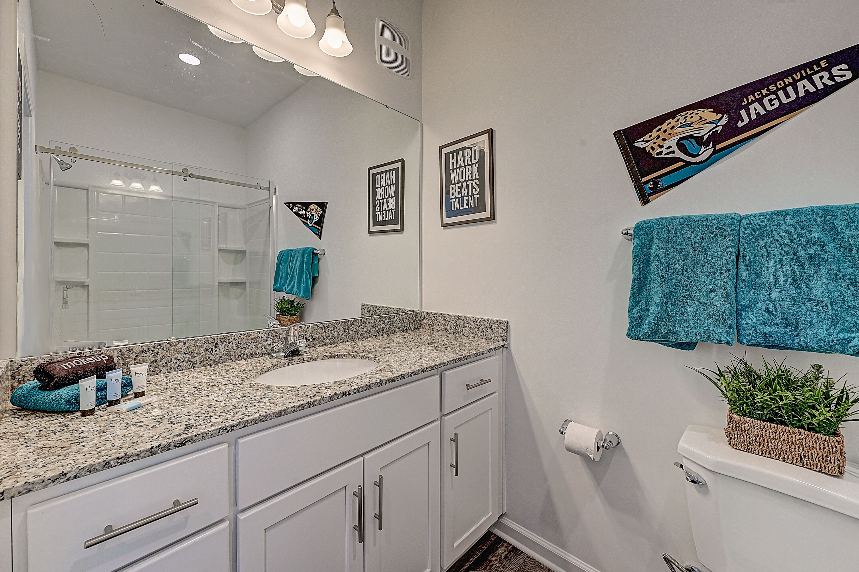 A bathroom with a white sink, granite countertop, and a mirror. There are towels hanging on the wall and a framed sign with a jaguar logo.