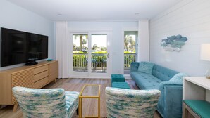 A modern living room with a large flat-screen TV, teal upholstered furniture, and a balcony with outdoor seating.