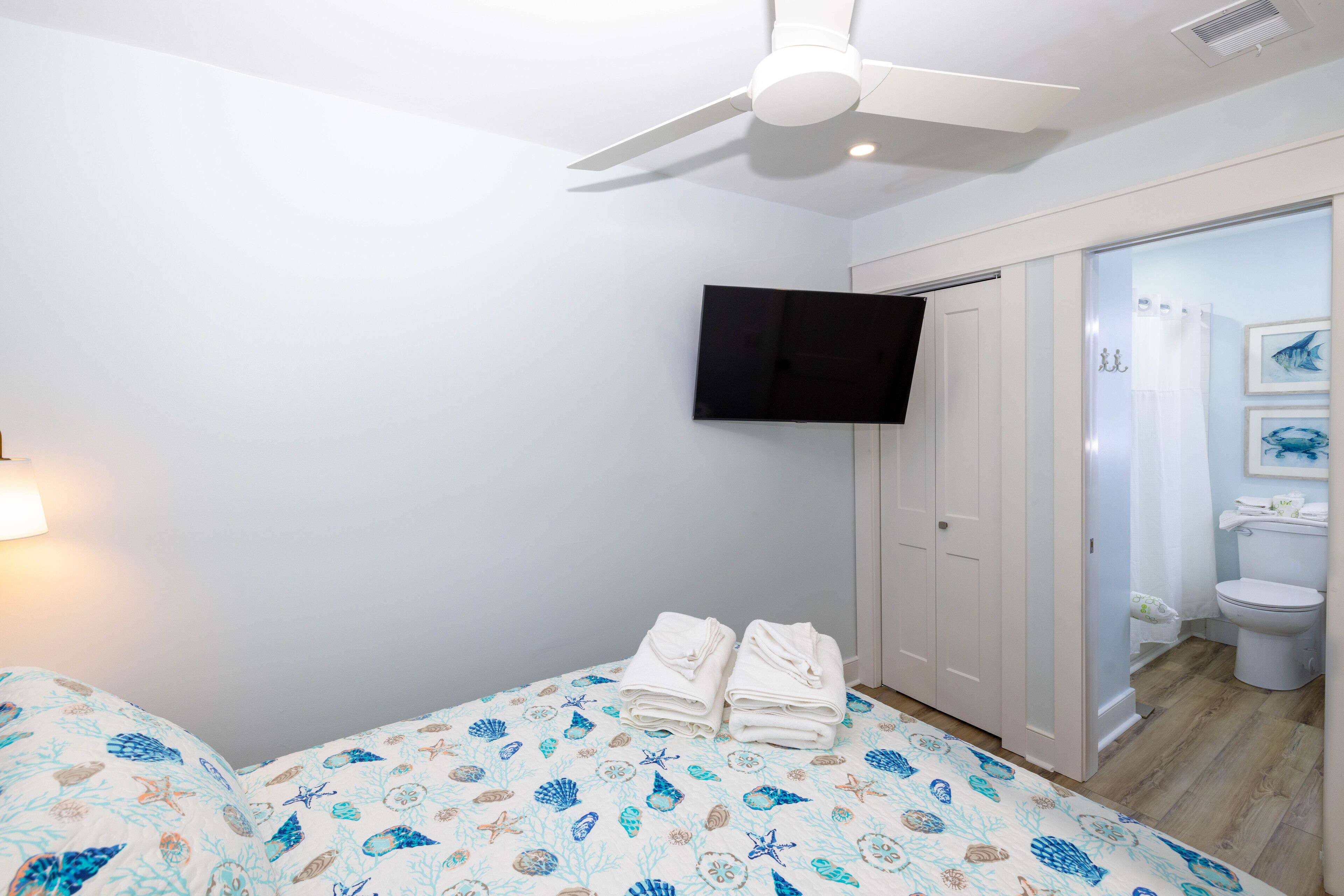 Un dormitorio con cama, ventilador de techo, televisor montado en la pared y baño visible a través de una puerta abierta.