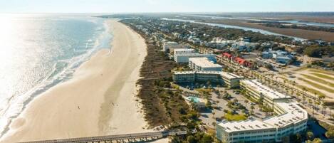 An aerial view of a coastal area with a beach, a bridge, and a hotel complex with multiple buildings and parking areas.
