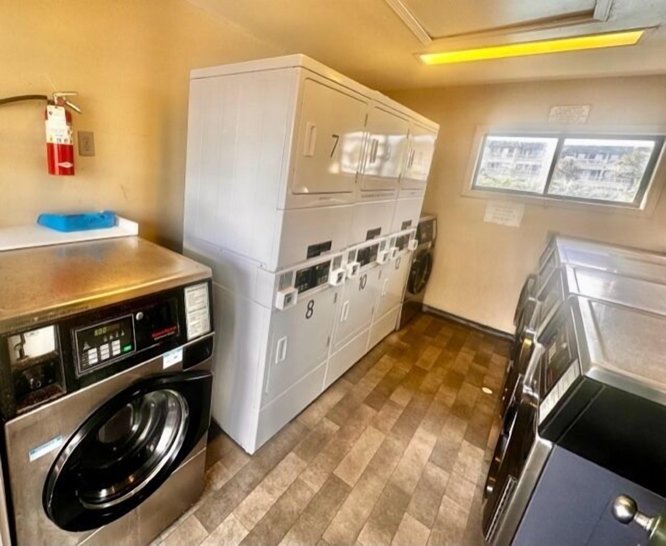 A laundry room with stacked washers and dryers, a fire extinguisher, and a view of the outside through a window.