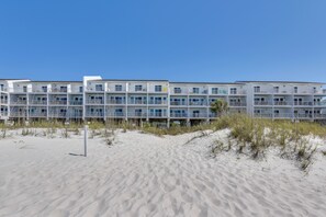Ein Hotel direkt am Strand mit mehreren Balkonen, ein sandiger Strand im Vordergrund und ein klarer, blauer Himmel.
