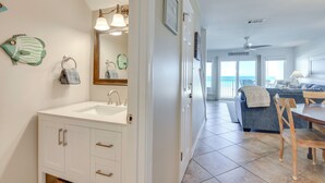 Un bagno con lavandino, specchio e porta asciugamani. C'è una zona pranzo con tavolo e sedie, e un soggiorno con divano e vista sull'oceano.