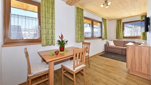 Une pièce avec un plafond en bois, une table à manger avec des chaises, un canapé, un téléviseur fixé au mur et de grandes fenêtres avec des rideaux verts.