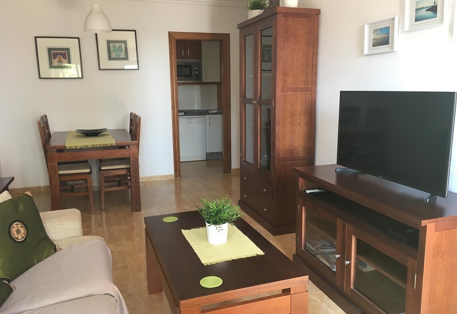 A living room with a sofa, a coffee table with a plant, a TV on a stand, a wooden cabinet, and a dining area with chairs and a table.