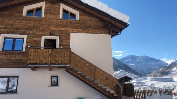 Una cabaña de madera con balcón, escaleras y vistas a montañas nevadas.