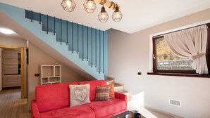 Una sala de estar con un sofá rojo, una mesita de centro, un libro, una escalera y una ventana con cortinas.