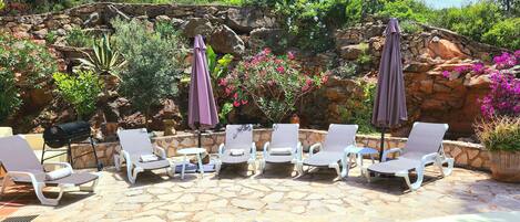 A poolside area with lounge chairs and umbrellas, surrounded by a stone wall and greenery.