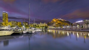 Un port de plaisance où sont amarrés des yachts, un bâtiment imposant dont le sommet est illuminé d’une lumière dorée, et un paysage urbain avec des bâtiments illuminés en arrière-plan.