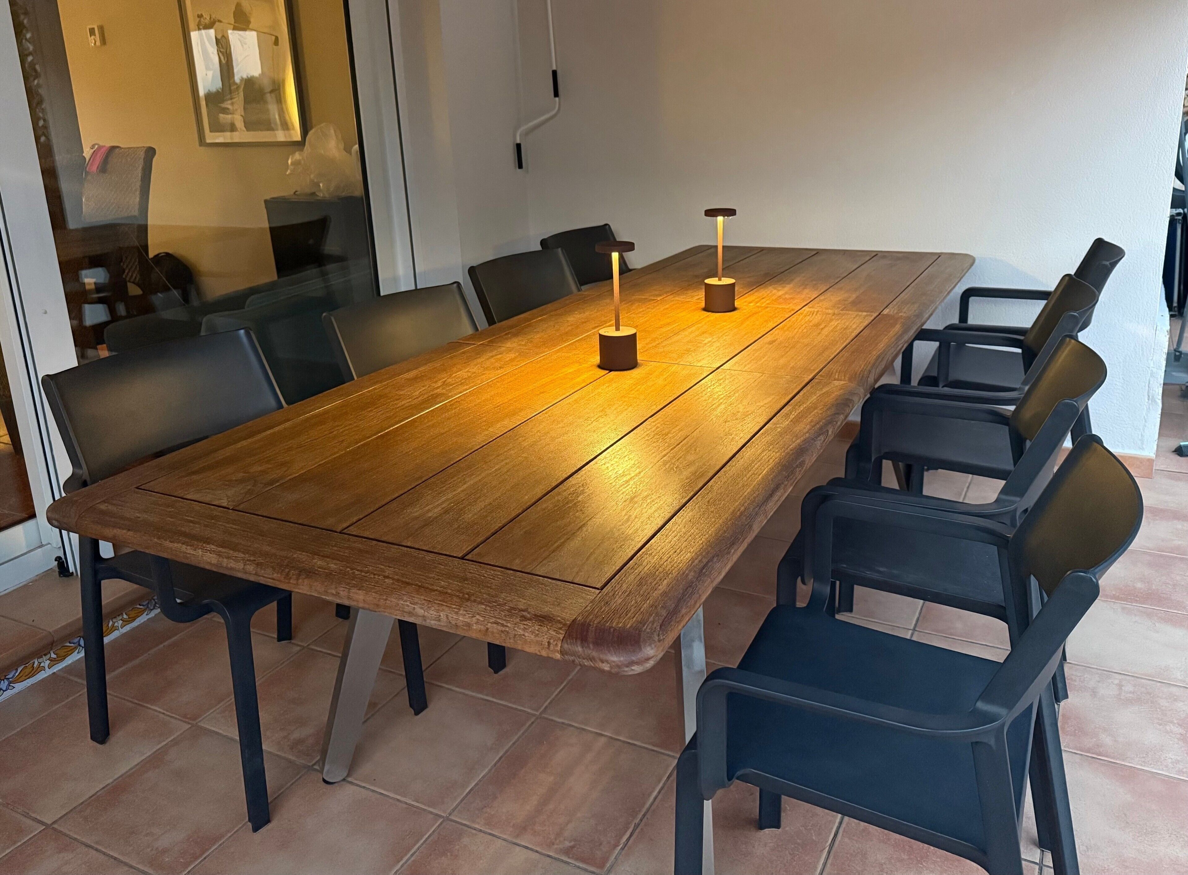 Una larga mesa de madera con sillas negras, dos lámparas colgantes y un separador de vidrio con un cuadro enmarcado.