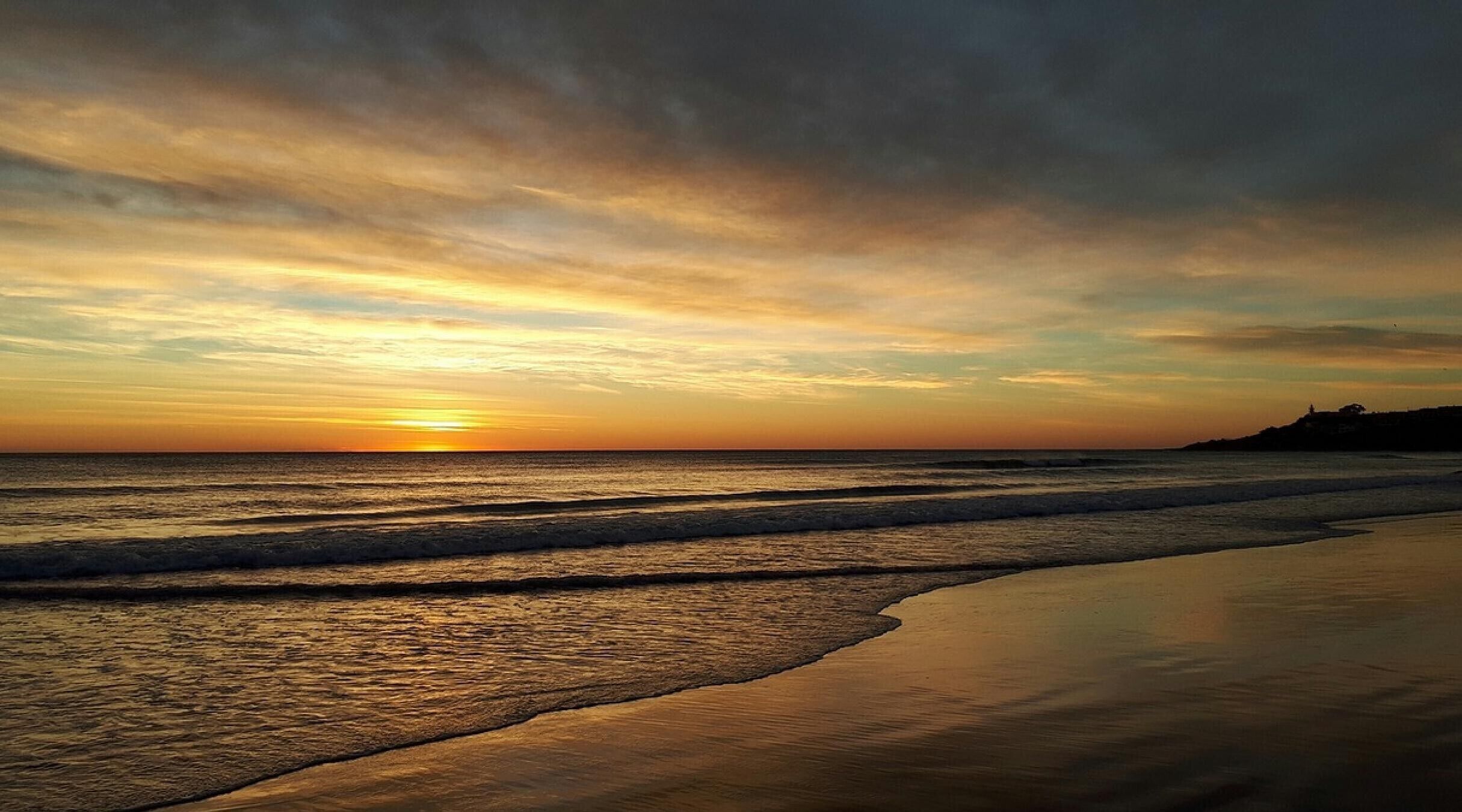 Una playa con olas, una puesta de sol y una costa distante.