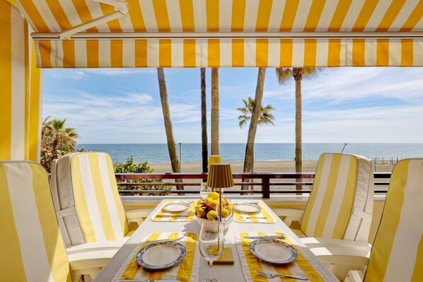A beachside table with yellow-striped chairs, a glass vase with flowers, and a view of the ocean.