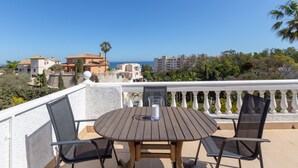 Une table ronde en bois, accompagnée de deux chaises, est placée sur un balcon qui donne sur une zone côtière, avec des bâtiments et des arbres.