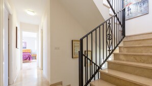 Un escalier avec des rampes noires menant à un palier, un couloir avec un lit au fond, et un tableau accroché au mur.