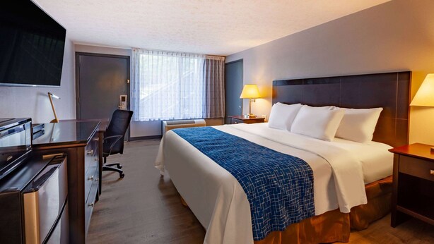 A hotel room with a bed, desk, chair, TV, and lamps. - Travelodge By Wyndham Charles Town/Harpers Ferry (Charles Town)