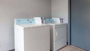 Two front-loading washers with control panels, a tiled floor, and a plain wall. - Travelodge By Wyndham Charles Town/Harpers Ferry (Charles Town)
