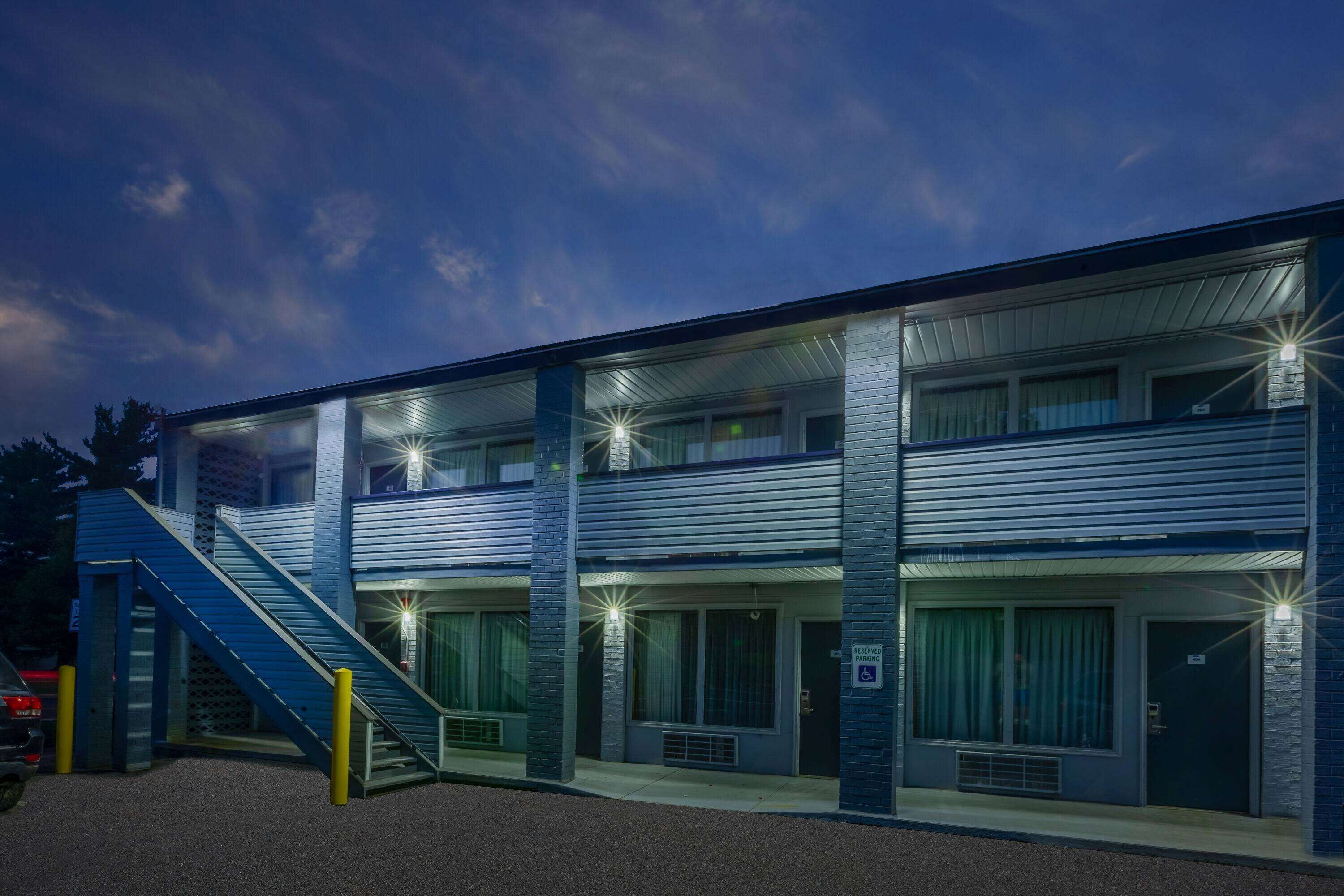 A multi-story motel with a staircase leading to the entrance, large windows, and a parking area.