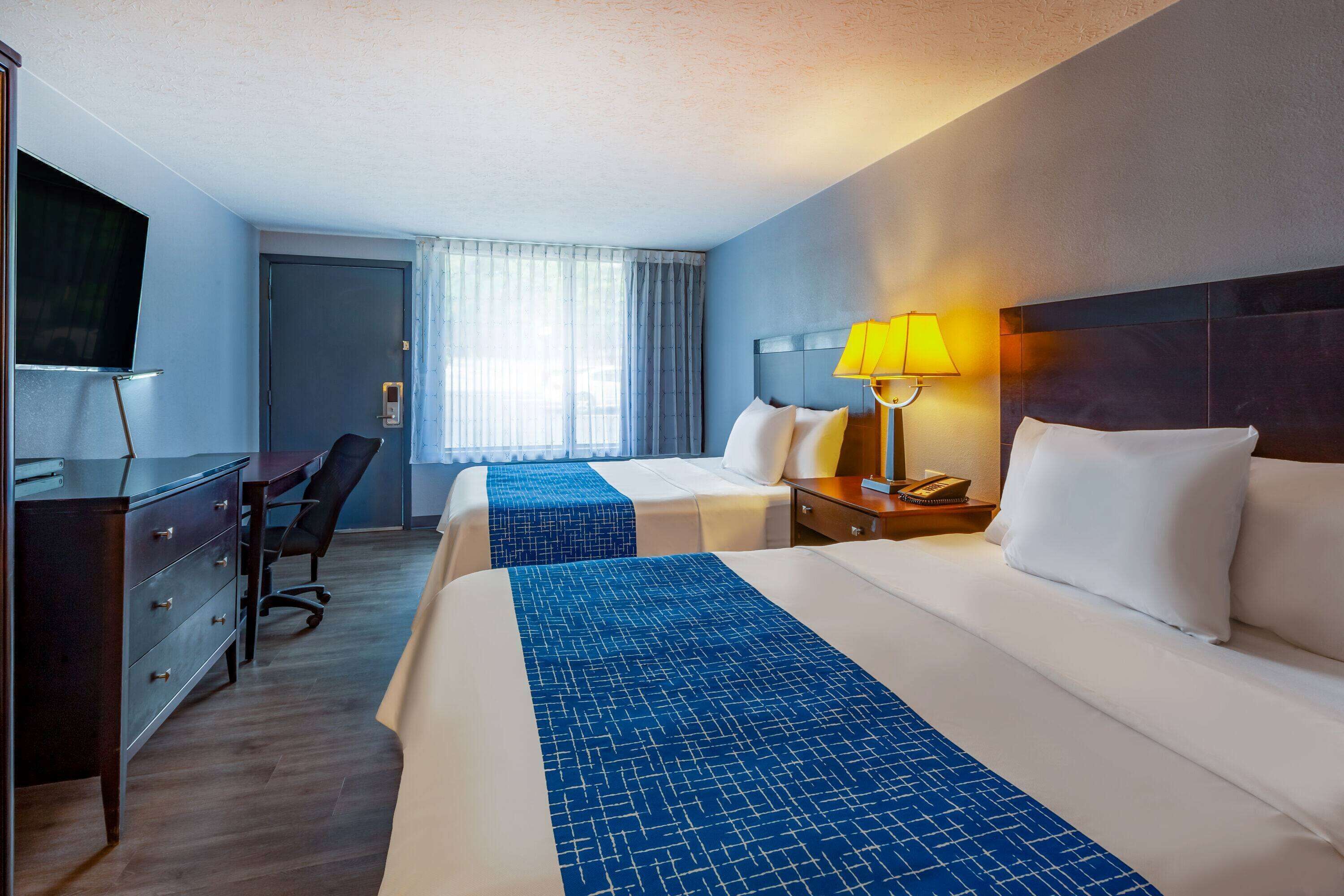 Quarto de hotel com duas camas, uma mesa, uma cadeira, televisão e janela com cortinas.
