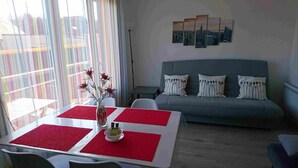 Un salon moderne avec un canapé gris, des sets de table rouges et blancs sur une table, et un vase contenant des fleurs séchées.