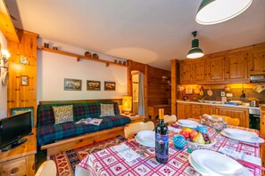 A cozy living room with a sofa, a dining table set for a meal, and a kitchen area with wooden cabinets. - Casa Cisi ski in-Ski out 20m (Livigno)