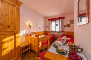 Ein gemütliches Zimmer mit einem Holzkasten, einem Esstisch mit rotem Tischläufer, einem Bett mit roter Bettwäsche und einem Fenster mit rotem Vorhang.