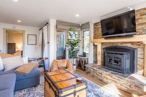 A cozy living room with a fireplace, a television mounted above it, and a large window with curtains. - Brand New Listing! 5 minute walk to Canyon Lodge! Newly Updated Two-Bed, Two-Bath, Mountainback #14 (Mammoth Lakes)