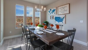 Une salle à manger avec une longue table, des chaises et un lustre. De grandes fenêtres offrent une vue sur un paysage désertique.