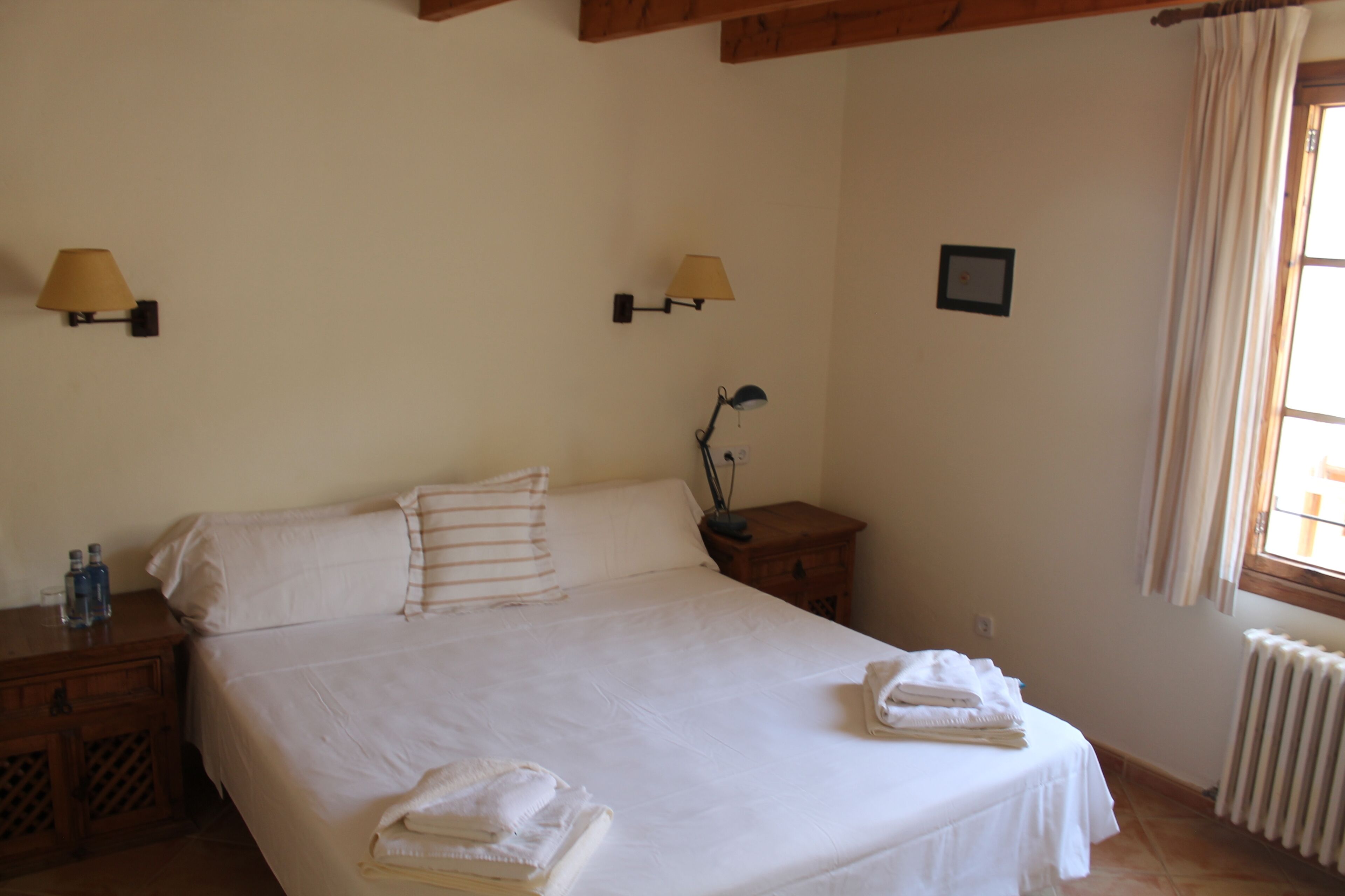 Una cama bien hecha con sábanas blancas, dos lámparas de noche, una mesita de noche con lámpara, un radiador y una ventana con cortinas.