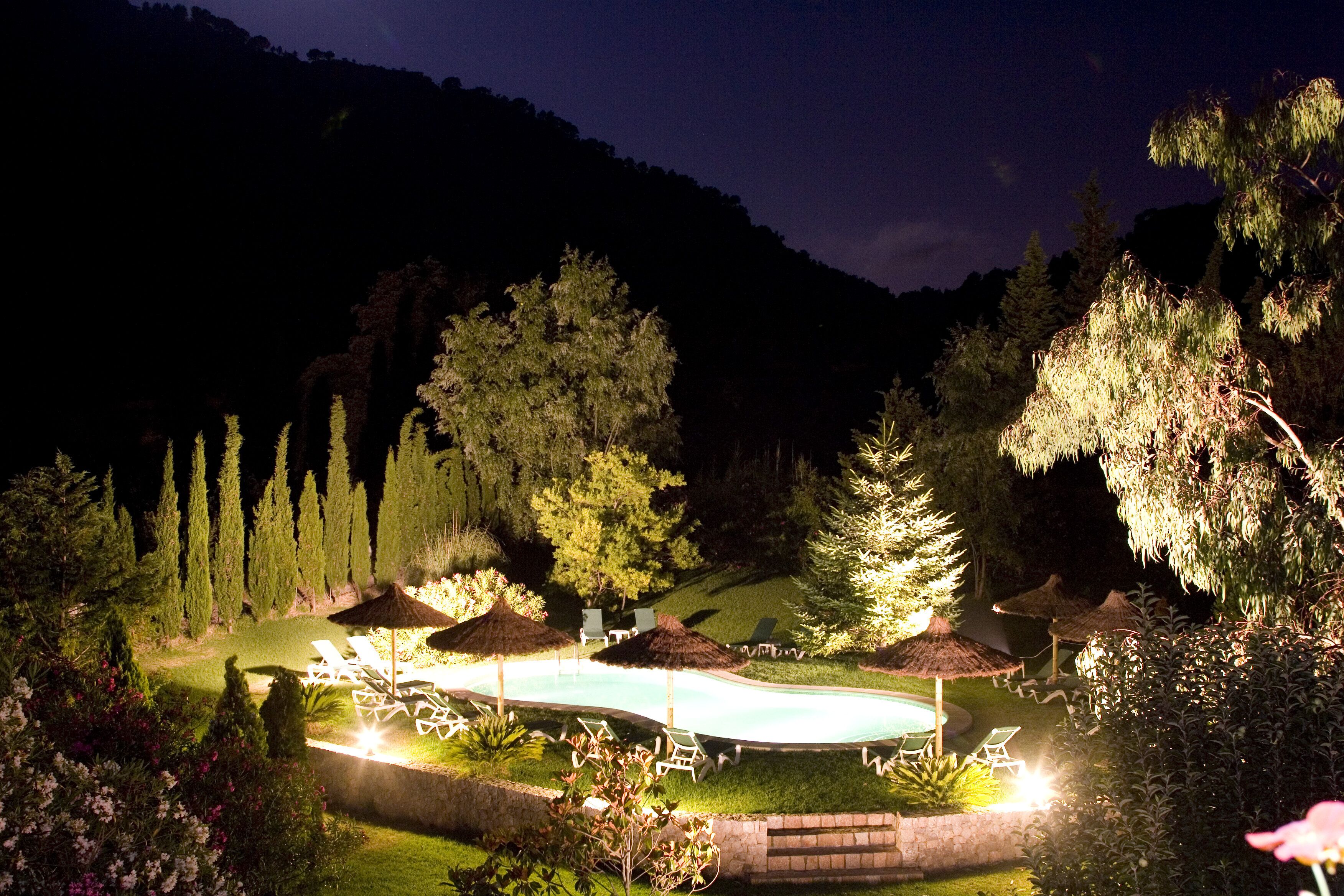 A nighttime view of a landscaped pool area with lounge chairs and umbrellas, surrounded by trees and hills.