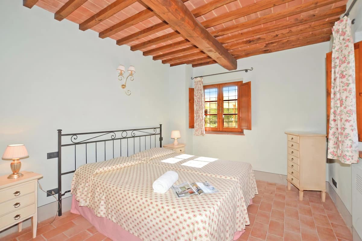 A bedroom with a bed, wooden ceiling beams, a window with curtains, a bedside table with a lamp, and a dresser.