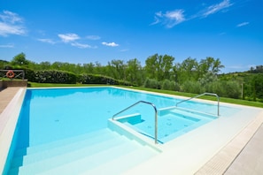 A rectangular swimming pool with clear blue water, surrounded by a paved deck and a grassy area with trees.
