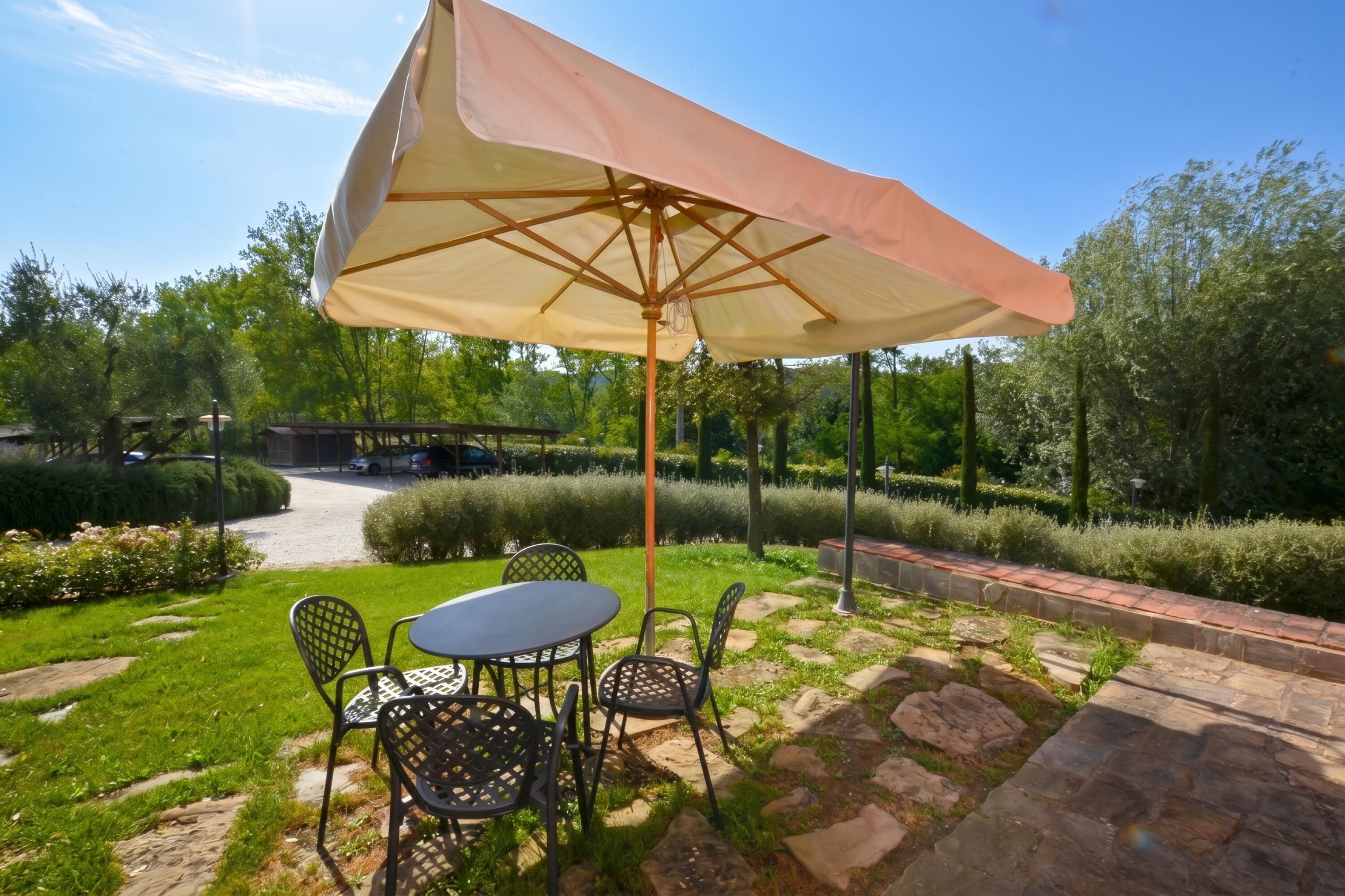 An outdoor patio with a table and chairs under a large umbrella, surrounded by greenery and a stone pathway.