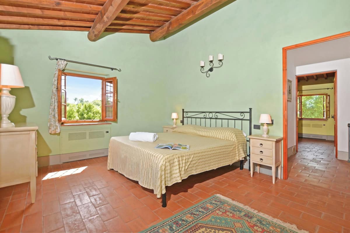 A bedroom with a bed, bedside tables, a window with curtains, a wall-mounted lamp, and a door leading to another room.