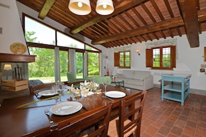Un coin repas avec une table en bois dressĂ©e pour deux personnes, un canapĂ© et une fenĂȘtre donnant sur un paysage verdoyant.